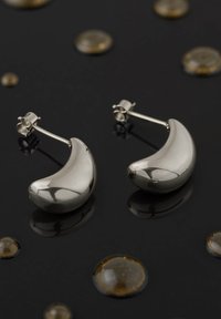 Orecchini in metallo color argento a forma di luna crescente, con una finitura lucida e chiusura posteriore, posizionati su una superficie nera riflettente.