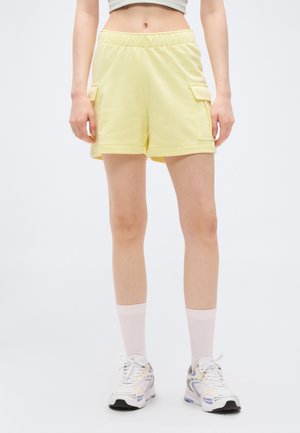 Personne portant un short cargo jaune clair, des chaussettes blanches montantes et des baskets blanches, debout devant un fond gris clair uni.