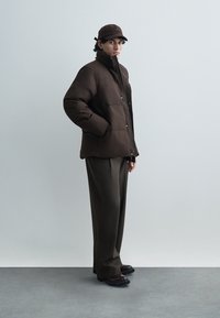 Brown puffer jacket with a high collar and snap buttons; paired with dark green trousers and black shoes; model wears a matching cap.