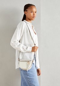 Bolso bandolera blanco de cuero suave con forma curva, detalles de hardware dorado y correa desmontable, combinado con una camisa de lino clara y denim azul.