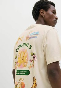 Krém színű, rövid ujjú póló, amelynek hátulján színes grafikák láthatók egy polipról, virágokról és egy pillangóról. A felirat: "ORIGINALS."