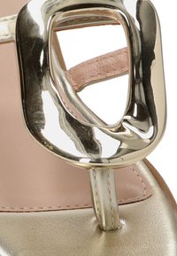 Acercamiento de una sandalia dorada con hebilla metálica gruesa y detalle de correa interna de cuero rosa.