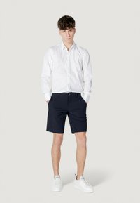 Camicia bianca a maniche lunghe con bottoni, pantaloni corti blu scuro e sneakers bianche. L'outfit presenta un design classico con una silhouette aderente.