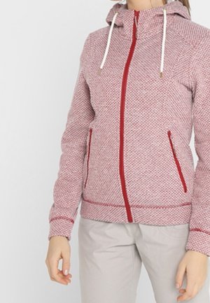 Femme portant un sweat à capuche rose texturé zippé avec une fermeture éclair rouge et des détails de poche, associé à un pantalon gris clair.