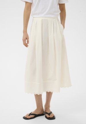 Persona vestida con una falda midi blanca plisada con dobladillo ondulado, camiseta blanca y sandalias negras tipo chancla, de pie frente a un fondo liso.