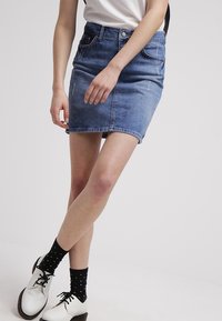 Denimkjol i blekt blå med en figurnära passform, framfickor och en lätt fransad nederkant. Buren med vita skor och svarta prickiga strumpor.