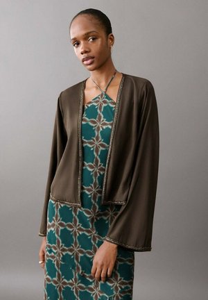 Mujer con un vestido halter de patrones en tonos verde azulado y marrón, y un cárdigan abierto de punto suelto en color oliva oscuro, sobre un fondo gris liso.