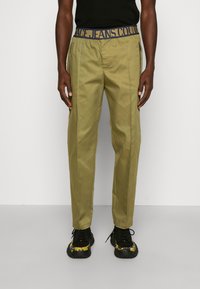 Man wearing olive green pants with elastic waistband and text, black shirt, and black sneakers with yellow floral design, standing.