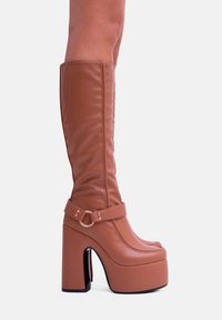 Stivali platform marroni in pelle sintetica al ginocchio con tacco chunky, caratterizzati da una cinghia con dettaglio a anello metallico. Texture liscia e punta arrotondata.