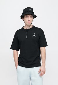 Svart bomulls-T-skjorte med rund hals og korte ermer, med en hvit Jordan-logo. Brukt med en matchende svart bøttehatt.
