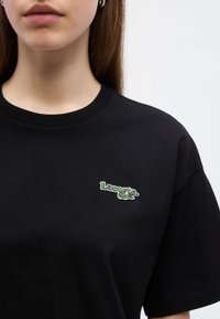 Γυναίκα που φορά μια μαύρη μπλούζα Lacoste με ένα μικρό πράσινο λογότυπο κροκόδειλου στο αριστερό στήθος, μπροστά σε λευκό φόντο.