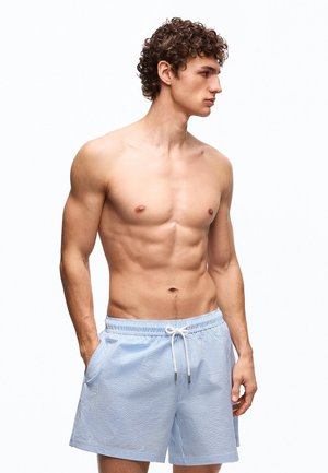 Ing nélküli fiatal férfi göndör hajjal, világoskék és fehér csíkos fürdőnadrágot visel, zsebében tartja a kezét, jobbra néz.