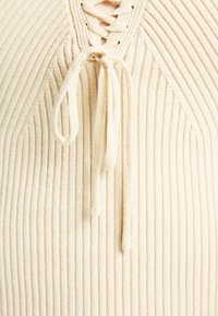 Krämfärgad ribbad tröja med snörning i halslinjen och dragsko-detalj. Mjuk textur med vertikala linjer genom tyget.