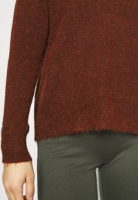 Pull doux couleur rouille avec un col rond côtelé et des manches longues. Légèrement court avec un ourlet brut, porté sur un pantalon vert olive.