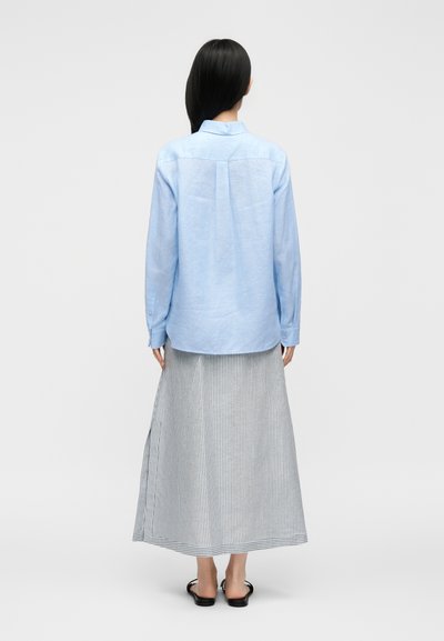 Femme debout, dos à la caméra, portant une chemise bleu clair à manches longues, une jupe rayée jusqu'aux chevilles et des sandales noires à bouts ouverts sur fond blanc.