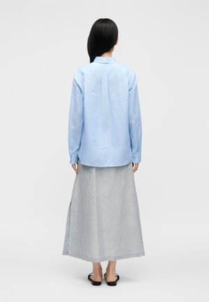 Žena stojaca chrbtom ku kamere, oblečená v svetlomodrú košeľu s dlhým rukávom, pruhovanú sukňu po členky a čierne sandále s otvorenou špičkou na bielom pozadí.