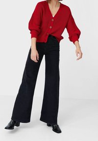 Cardigan rouge en tricot avec un col en V, orné de trois grands boutons, associé à un jean taille haute noir à jambes larges et des chaussures noires à talons blocs.