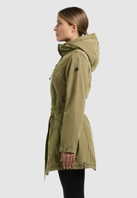 Ženska stoji v profilu, oblečena v olivno zeleno kapuco parko plašč in črne hlače na enobarvni podlagi.