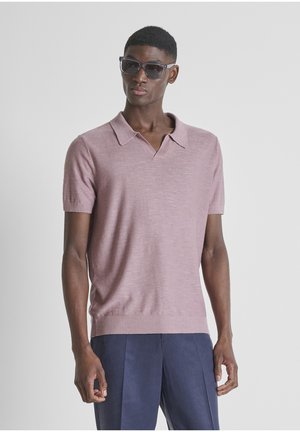 Man met donkere zonnebril, een lichtpaarse katoenen korte mouw polo en donkerblauwe broek, staand tegen een effen achtergrond.