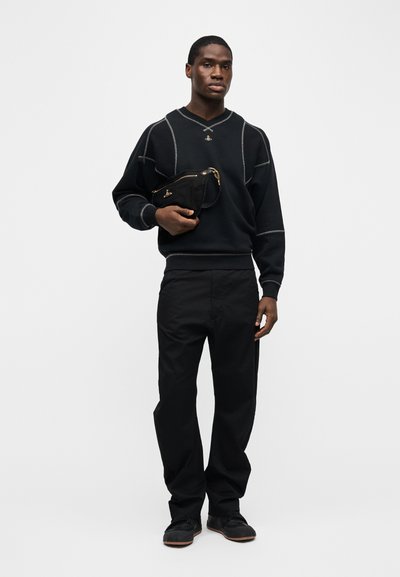 Άνδρας που φοράει μαύρο φούτερ με λευκές ραφές, μαύρο παντελόνι και μαύρα παπούτσια, κρατώντας μια μαύρη θήκη με χρυστό φερμουάρ πάνω σε λευκό φόντο.