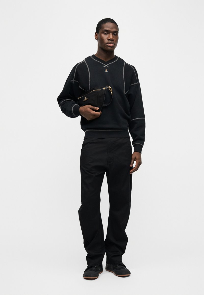 Uomo che indossa una felpa nera con cuciture bianche, pantaloni neri e scarpe nere, che tiene una borsa nera con cerniera dorata su sfondo bianco.