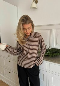 Prugasta, dugorukava majica u taupe i tamnosmeđoj boji, s ovratnikom i detaljem dugmadi. Nosena uz crne hlače, drži bijelu šalicu.