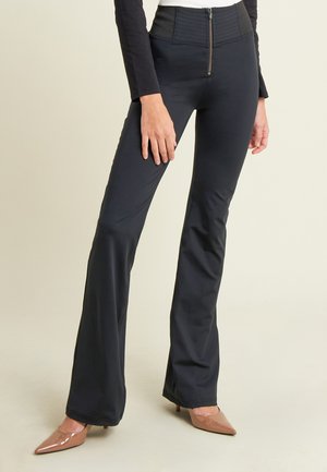 Donna che indossa pantaloni neri a vita alta, a palazzo, con zip frontale e décolleté beige a punta, in piedi contro uno sfondo neutro.