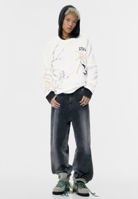 Sweatshirt alb cu imprimeuri grafice și accente gri, purtat cu blugi baggy de culoare închisă și încălțăminte gri cu șireturi verzi.