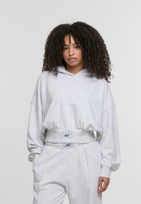Hoodie corto grigio chiaro con cappuccio regolabile, maniche ampie e orlo a coste. Abbinato a pantaloni della tuta coordinati con toppe con logo.