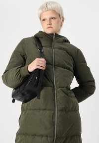 Geacă puf verde cu fermoar, cu glugă și textură cusută. O geantă crossbody din piele neagră stă în mâna modelului.