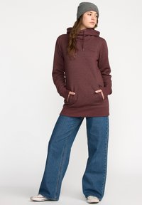 Sweatshirt bordeaux avec un col cheminé et des poches avant, associé à un jean large bleu et un bonnet en tricot gris. Texture lisse, design décontracté.
