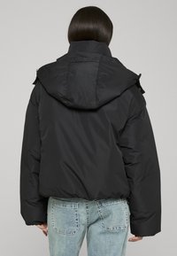 Svart vattert jakke med hette, med en kort design og elastisk nederkant. Brukt over lyseblå jeans med dekorative sømmer.