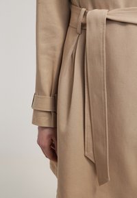 Gabardina beige con cinturón atado, que presenta una tela estructurada y texturizada, y puños doblados. Primer plano del detalle de la cintura y la manga.