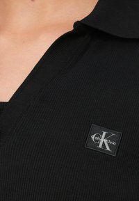 Schwarzes, geripptes Poloshirt mit einem Logo-Patch "Calvin Klein" und einem auffälligen "CK"-Design auf der linken Seite des Kragens.