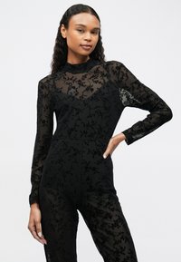 Salopetă neagră cu mâneci lungi, din materiale transparente, având un design din dantelă florală. Guler înalt, bust ajustat și picioare drepte. Material elegant și ușor.
