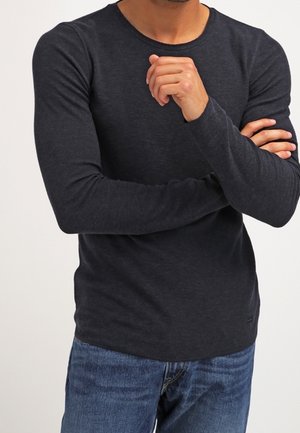 Maglietta a maniche lunghe e collo tondo di colore grigio scuro, realizzata in tessuto morbido. Design aderente con dettagli di cucitura minimali, abbinata a jeans blu.