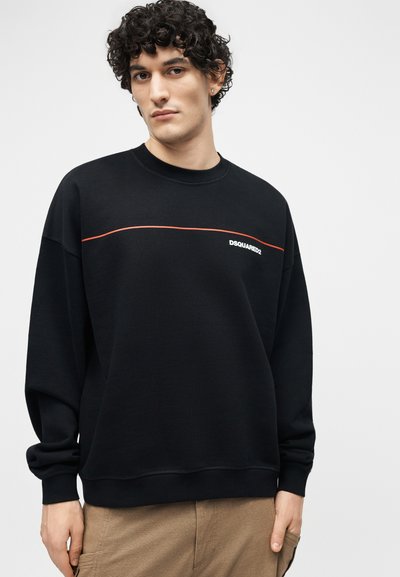 Νεαρός με σγουρά μαλλιά που φοράει μαύρο φούτερ DSQUARED2 με λεπτή κόκκινη γραμμή κατά μήκος του στήθους και μπεζ παντελόνια.