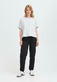Kvinne som står iført en løs lys grå t-skjorte med kort erm, svarte joggebukser og hvite joggesko med svarte striper, på en enkel lys bakgrunn.