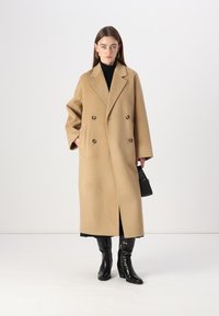 Cappotto in lana color cammello doppiopetto con revers oversize. Presenta due tasche anteriori ed è indossato sopra un dolcevita nero e un vestito.