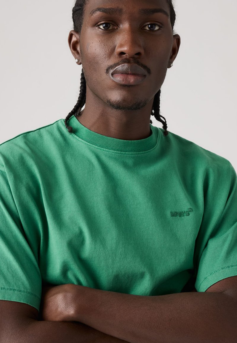 Jeune homme aux cheveux tressés et boucles d'oreilles, portant un t-shirt vert Levi's, bras croisés, expression neutre, arrière-plan clair uni.