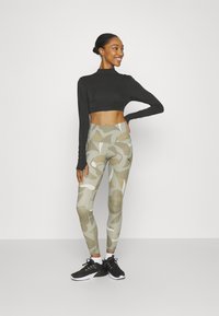 Top negro de manga larga cortado, combinado con leggings de estampado de camuflaje en beige, crema y oliva. Zapatillas deportivas negras completan el conjunto.