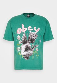 Zelené bavlněné tričko s černobílým motivem cheruba mezi květinovými vzory, s nápisem "obey" vytištěným růžovou barvou nad ním.