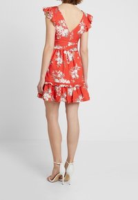 Červené květinové mini šaty s volánovými rukávy, V-výstřihem, stahovaným pasem a vícerečnými sukněmi. Oblečeno s bílými sandály na vysokém podpatku.