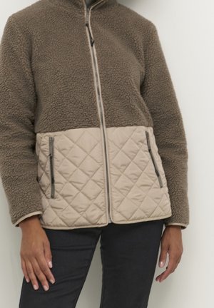 Person iført en brun fleecejakke med beige quiltet bund og lynlåslommer, kombineret med mørke bukser, stående mod en hvid baggrund.