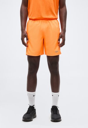 Mann trägt knallorange Nike-Shorts, weiße Nike-Crew-Socken und schwarze Sportschuhe, steht vor einem einfarbigen Hintergrund.