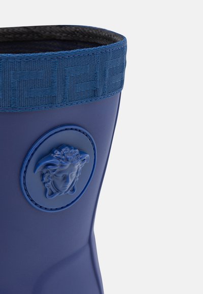 Botte en caoutchouc bleue avec une bande texturée bleue en haut. Présente un logo circulaire de Méduse et une surface lisse et brillante.