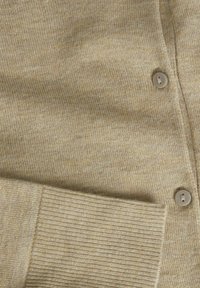 Virkattu beige kardigan pehmeällä tekstuurilla, jossa on ribbejä hihassa ja neljä pyöreää, vaaleaa nappia edessä.