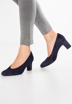 Talons en daim bleu marine avec un bout rond et un talon bloc de 5 cm, portés avec des leggings gris jusqu'à la cheville. Texture lisse et design simple.