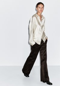 Kremowa satynowa koszula z guzikami w luźnym fasonie, zestawiona z ciemnobrązowymi spodniami z korduroju o szerokich nogawkach i czarnymi botkami za kostkę.