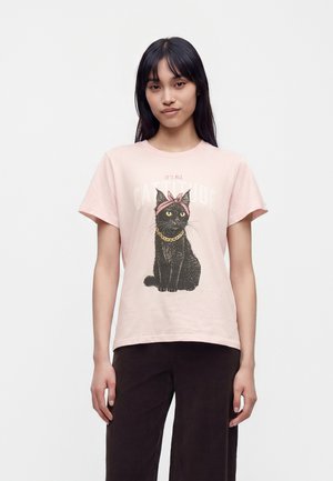 Jeune femme aux longs cheveux foncés portant un t-shirt rose clair avec une illustration d’un chat noir avec un bandeau rose et une chaîne en or.